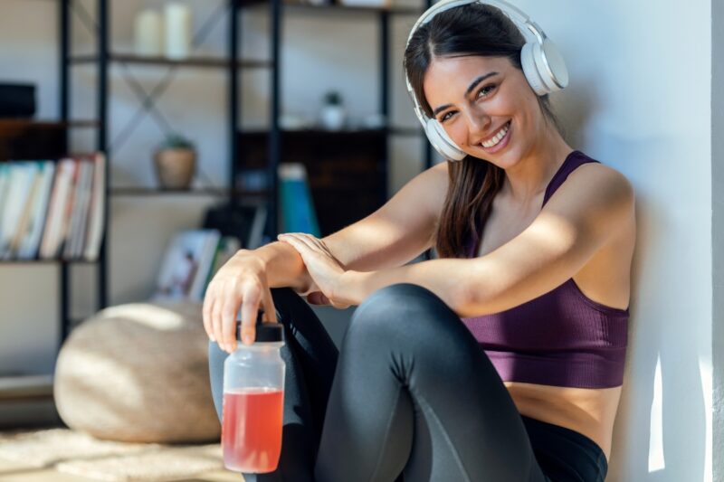 devojka se smeje, drži slušalice na ušima i pije pre workout 